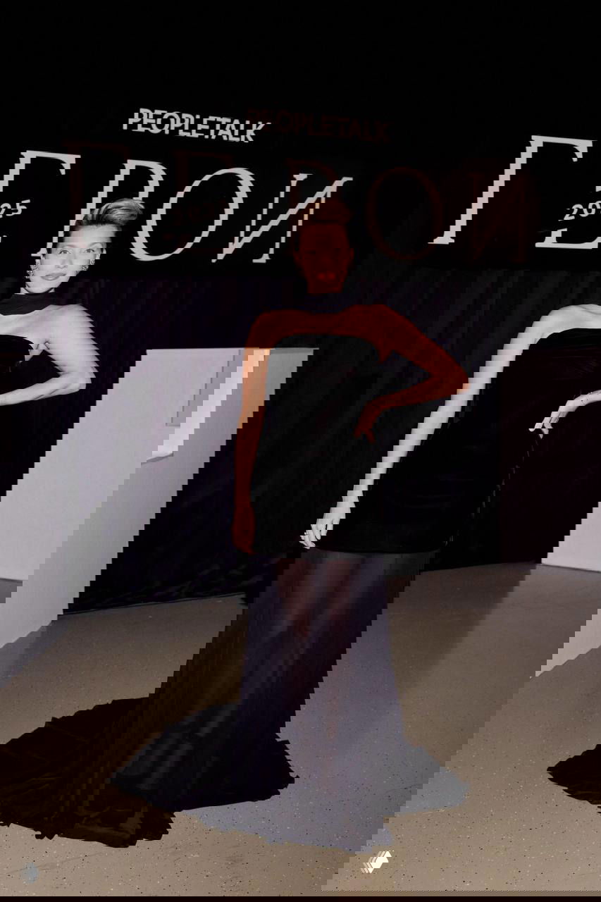 Женщина в открытом чёрном платье с прозрачной юбкой позирует на фоне логотипа «Герои 2025» — элегантный вечерний look для гала-вечера.