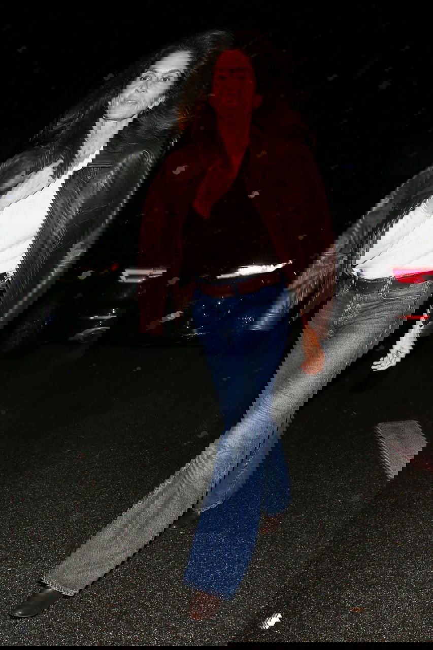 Папарацци‑кадр в полный рост: женщина в коричневой куртке и джинсах шагает по мокрому асфальту вечером; видны дождевые капли и отражения.