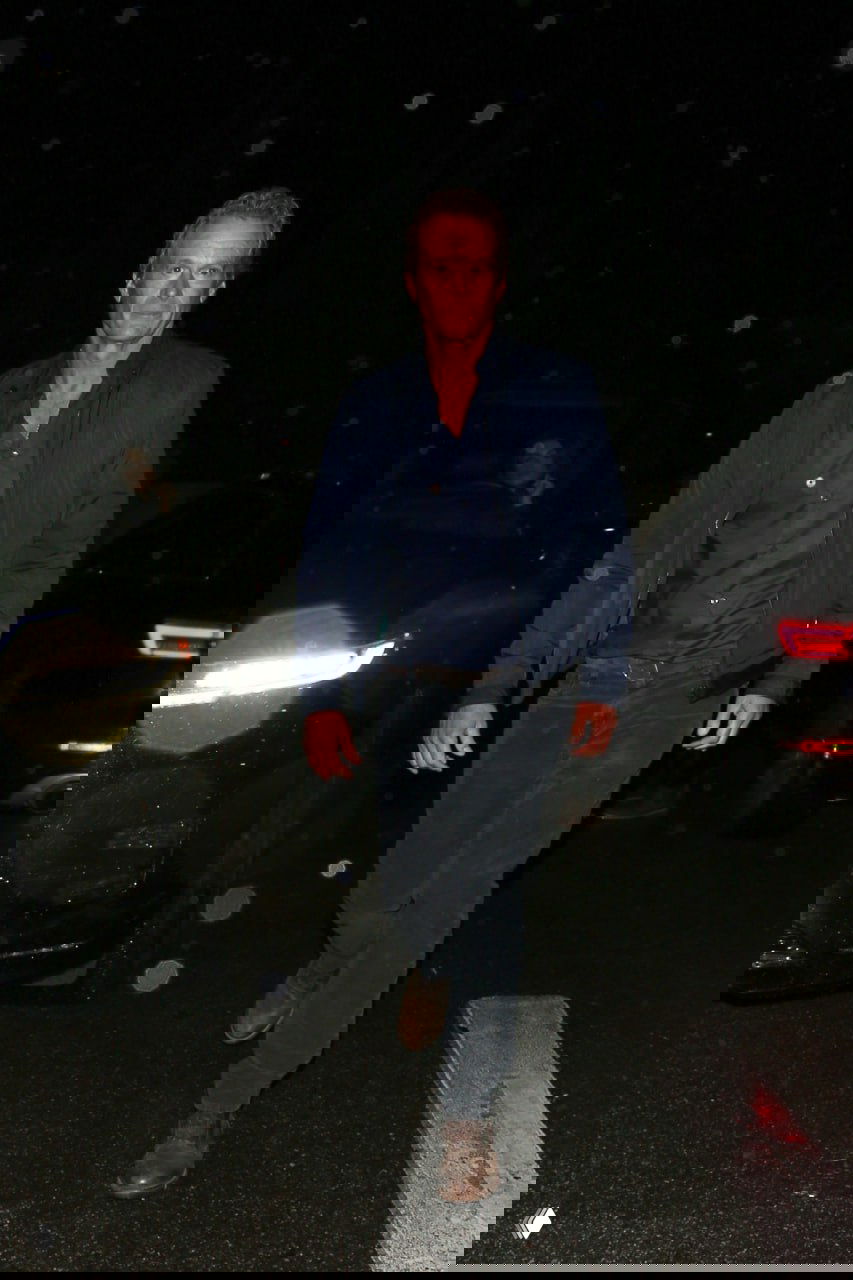 Папарацци‑снимок: мужчина в тёмной куртке и джинсах идёт по парковке ночью; фон — автомобили, уличные огни и блики на объективе.