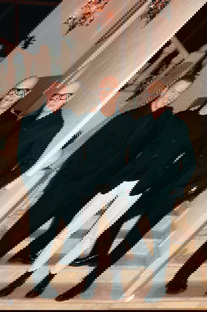 Трое мужчин в вечерних костюмах позируют на мраморной лестнице в торжественном интерьере — кадр с благотворительного вечера, праздничная атмосфера.