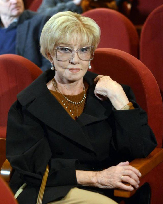 Фото пожилой женщины в зрительном зале, изображение сильно искажено цифровыми артефактами и цветовыми сдвигами, видно кресла в аудитории.