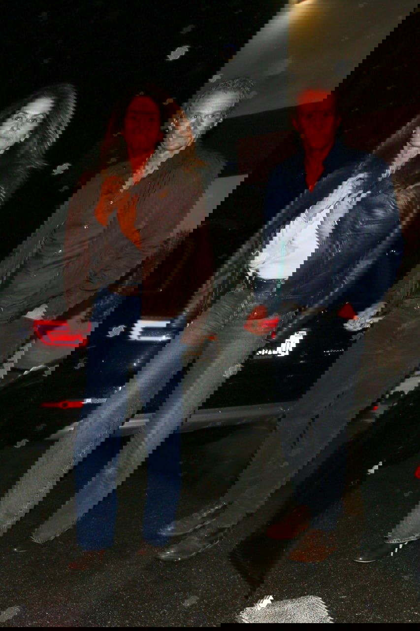 Папарацци‑снимок: женщина в коричневой кожаной куртке и джинсах идёт по мокрой улице; вокруг видны вспышки фотоаппаратов и блики.