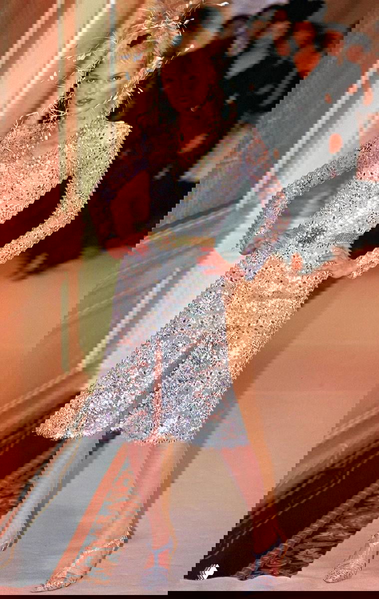 Кейт Мосс на подиуме в блестящем платье и с выразительной постановкой руки, показ высокой моды с гламурным образом и сценическим освещением.