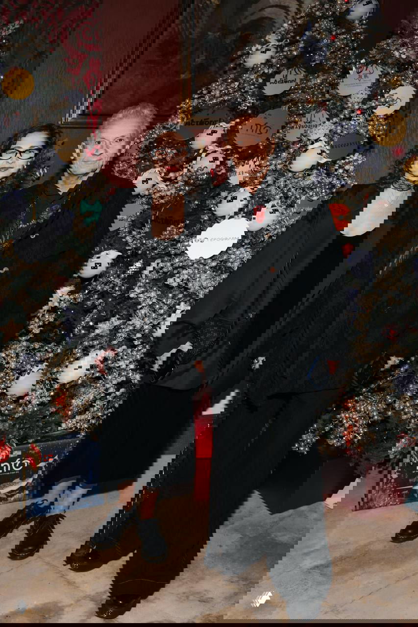 Пара в тёмной одежде стоит на фоне украшенной рождественской ёлки и праздничного декора — фотография из фойе благотворительного вечера.