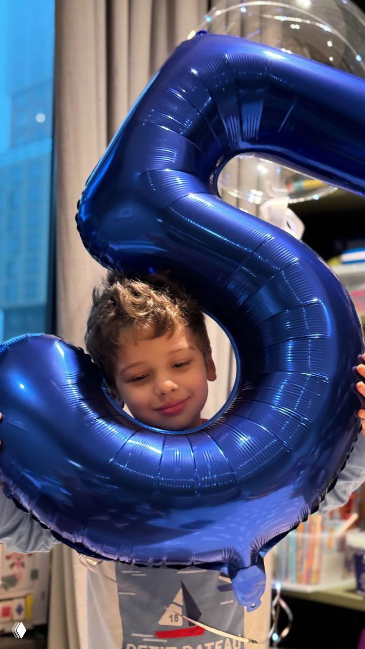 Маленький Гаспар улыбается и держит большой синий фольгированный шар в форме цифры «5» на домашнем празднике, праздничная атмосфера.