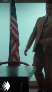 Мужчина в деловом костюме с пистолетом у стола на фоне американского флага — кадр в стиле шпионского триллера, атмосфера 70‑х годов.