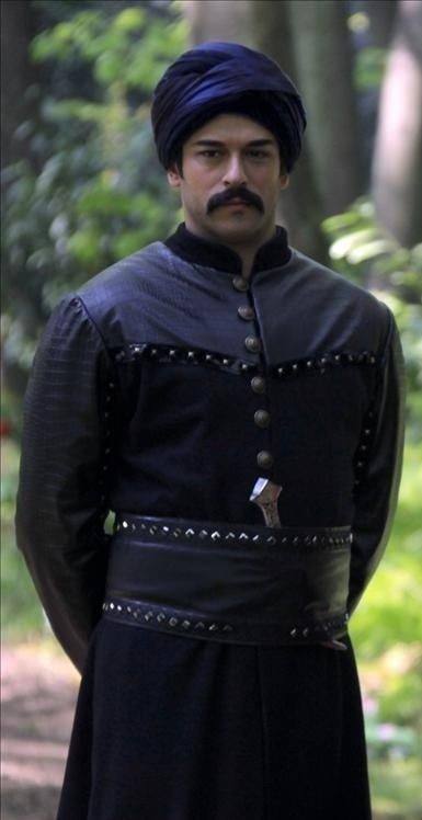 Мужчина в историческом костюме с тюрбаном стоит в лесной чаще — атмосферный кадр со сценическими деталями, костюмной отделкой и природным фоном.