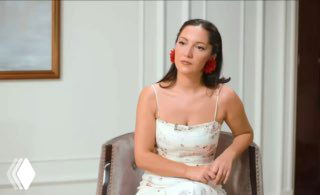 Кадр интервью: сидящая женщина в светлом платье в кресле, нейтральный студийный фон, иллюстрация дискуссии о роли женщин