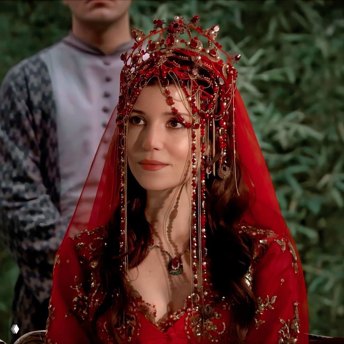 Крупный план Хюррем в красном головном уборе и богатых украшениях: прозрачная вуаль, декоративные детали и спокойное лицо героини в кадре.