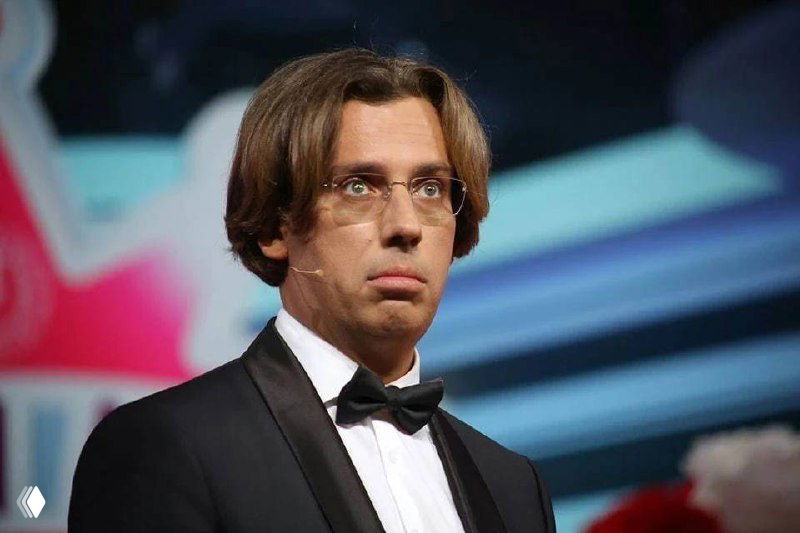 Мужчина на сцене в смокинге и очках, с микрофонной гарнитурой, внимательно смотрит вперёд; размытый сценический фон и свет