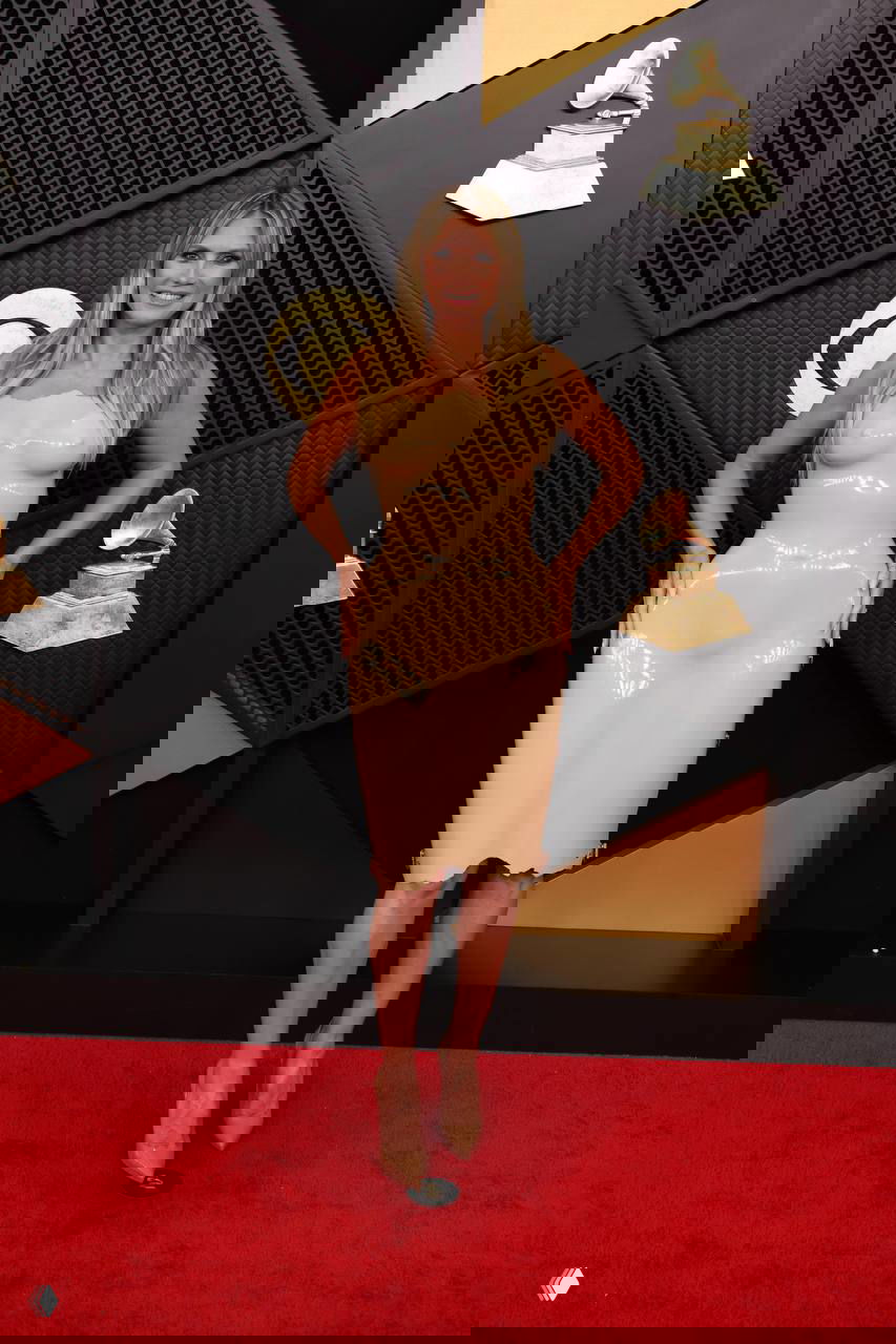 Хайди Клум позирует у фотозоны «Грэмми — 2026» в откровенном телесном платье, вид спереди, улыбка, золотые граммофоны и чёрный фон фотозоны.