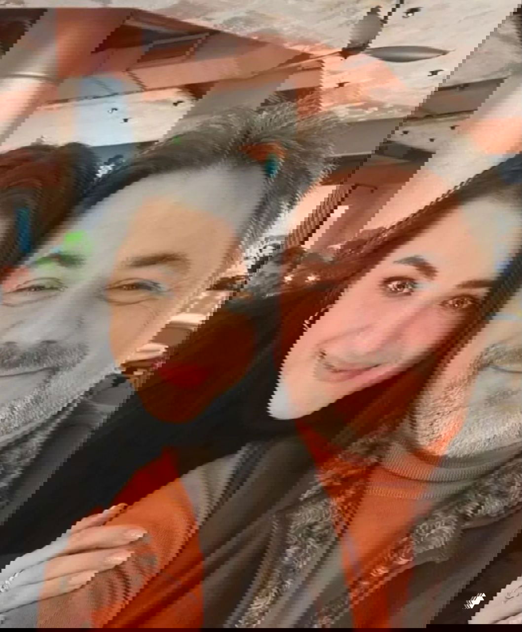 Женщина в пальто улыбается в бытовой обстановке — постановочный кадр с декором интерьера на заднем плане.