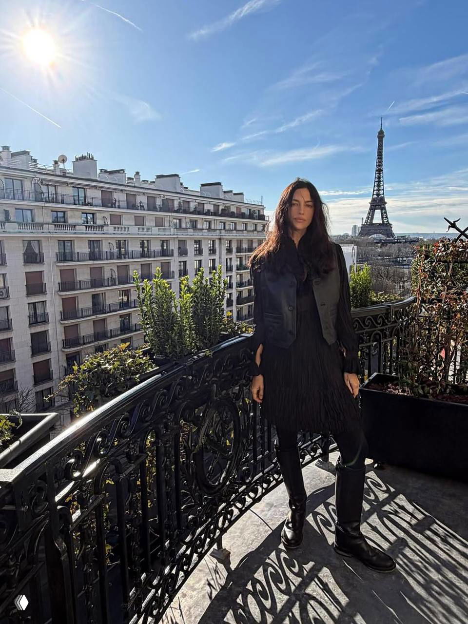 Полноформатный снимок Ирины Шейк на балконе в черном платье и ботинках на фоне парижской архитектуры и Эйфелевой башни днем.