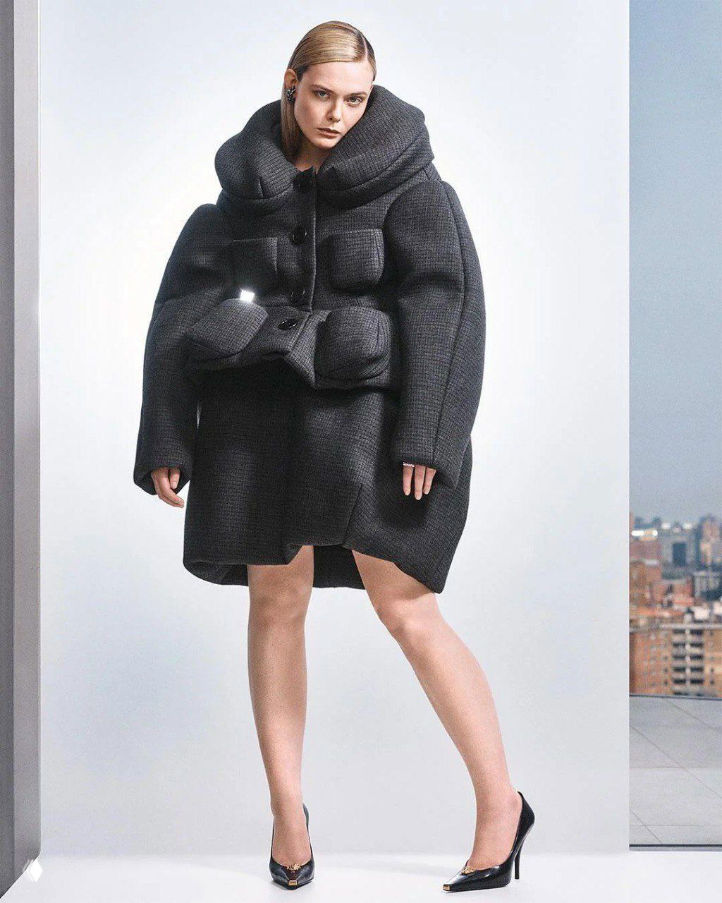 Полноформатный портрет в объемном стеганом пальто на нейтральном фоне с намеком на городской пейзаж — стилизованный образ из фотосета для журнала.