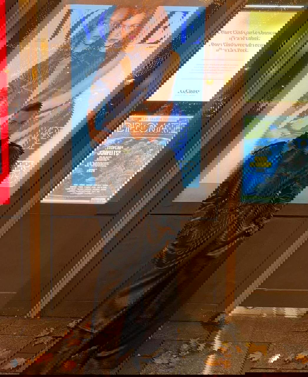 Ночной уличный снимок: Ирина в длинном черном кожаном пальто позирует у витрины с обложкой Vogue и рекламными плакатами позади.