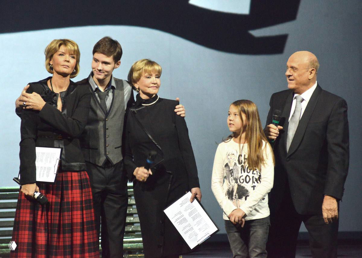 Группа людей на сцене во время церемонии: несколько взрослых и ребёнок держат микрофоны и листы текста, театральный фон позади них.