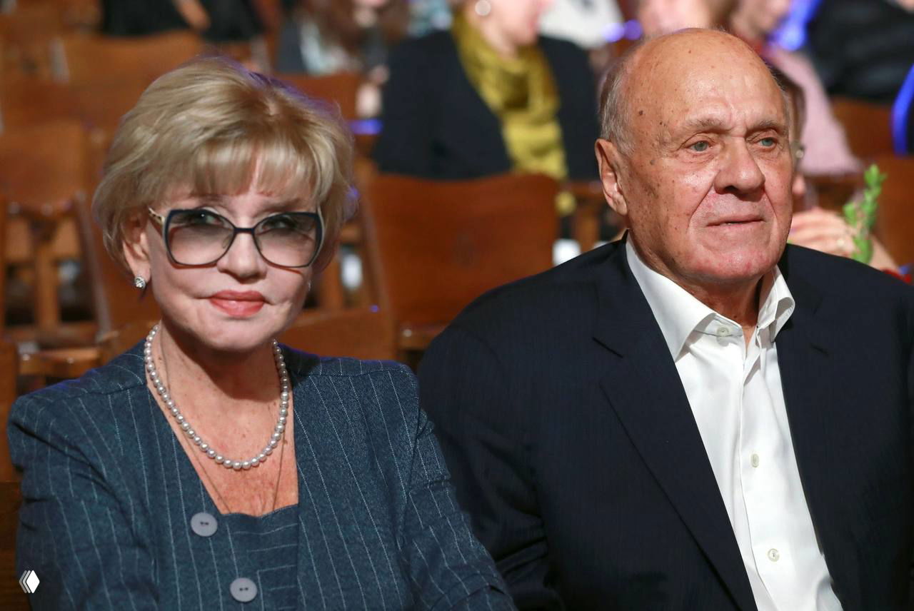 Пара пожилых людей в зрительном зале: женщина в очках и украшениях и мужчина в тёмном костюме, вокруг видны другие зрители и кресла.