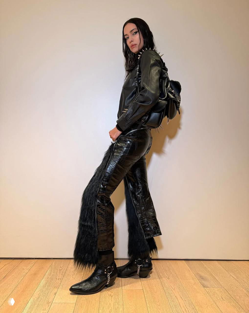 Женщина в чёрном кожаном комплекте позирует в полный рост на нейтральном фоне; акцент на текстуре кожи, ботинках и меховых вставках на брюках.