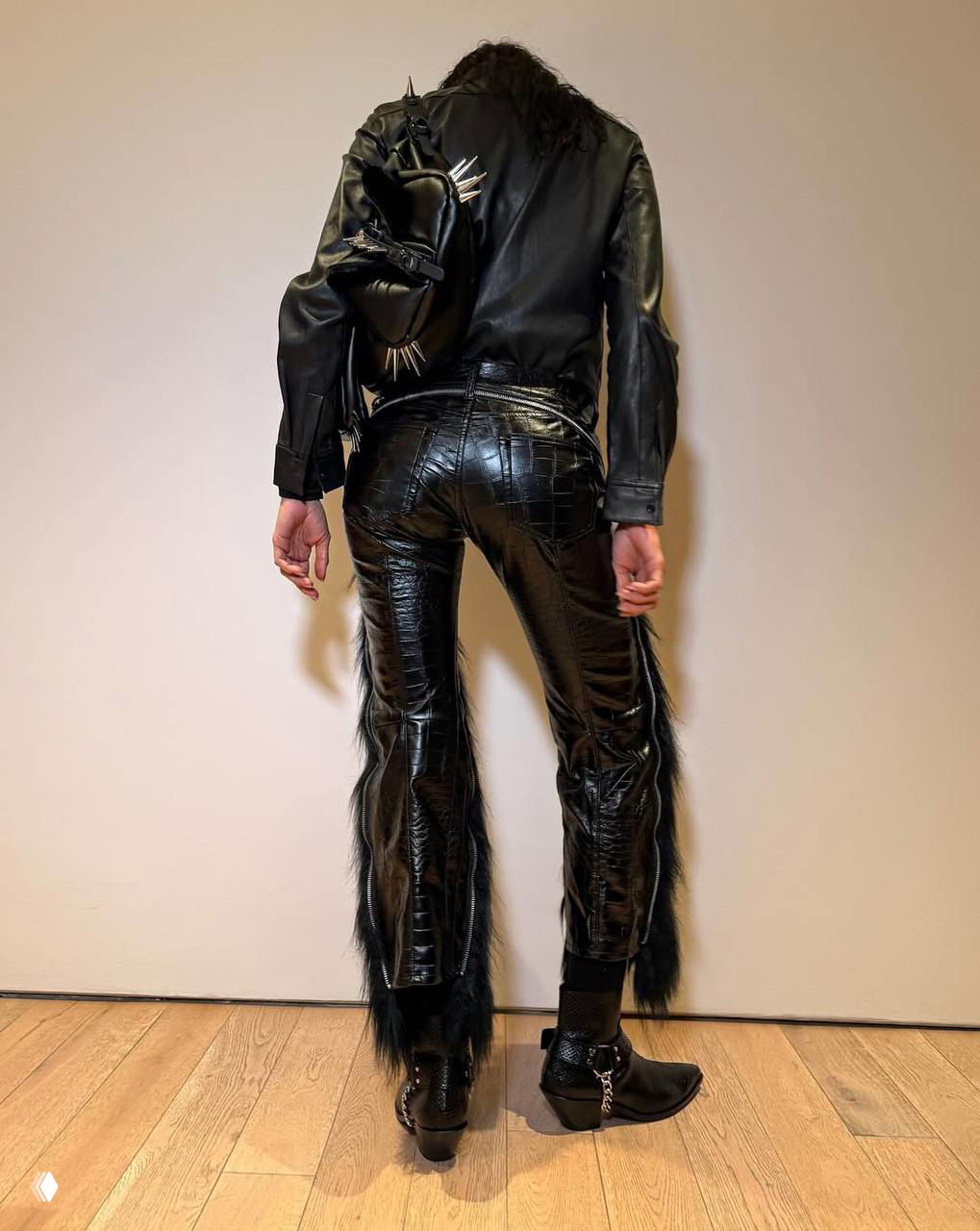 Задний план: женщина в кожаной рубашке и кожеобразных брюках снята со спины; видно фактуру ткани и детали кроя на нейтральном фоне.