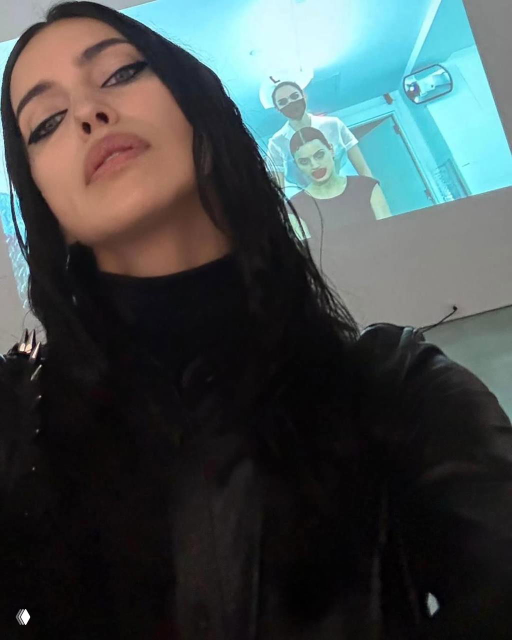 Крупный план лица женщины в тёмном макияже на фоне проекции изображения; акцент на стрелках и глянцевом тоне кожи и волос.