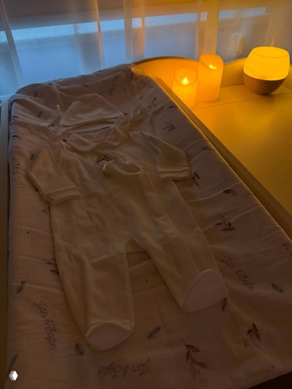 Белый человечек и пелёнальный стол с мягким светом и свечами: одежда для новорождённого на фоне палаты после родов.