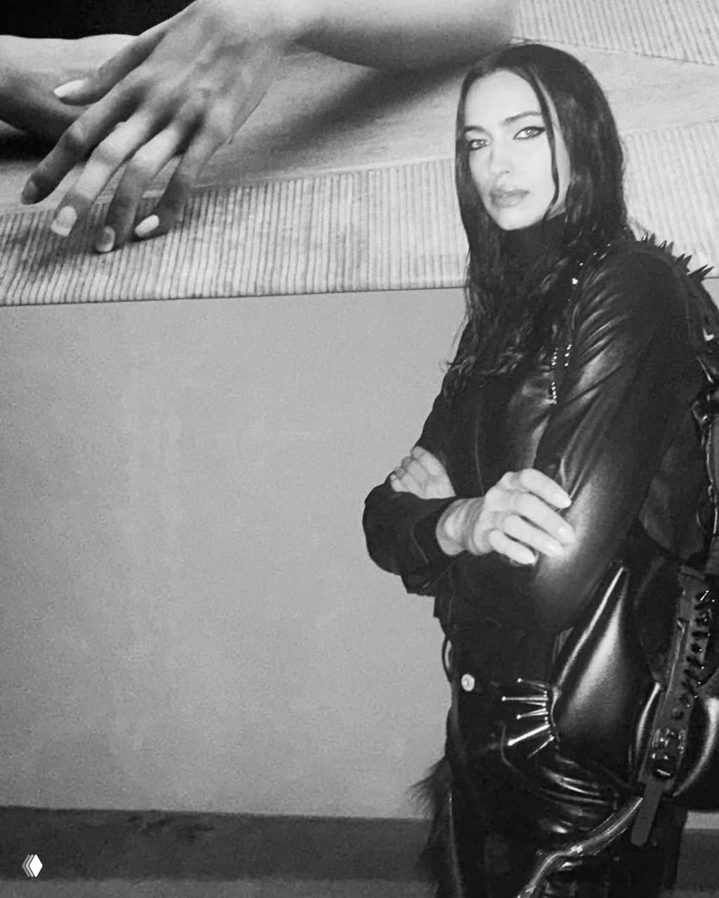 Чёрно‑белый портрет женщины в кожаном образе на фоне художественного полотна; снимок подчёркивает контраст текстур и позу с перекрещенными руками.