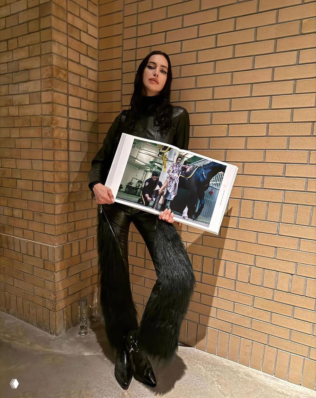 Женщина в кожаном костюме стоит у кирпичной стены, держит разворот журнала; видны длинные меховые детали брюк и черные остроносые ботинки.