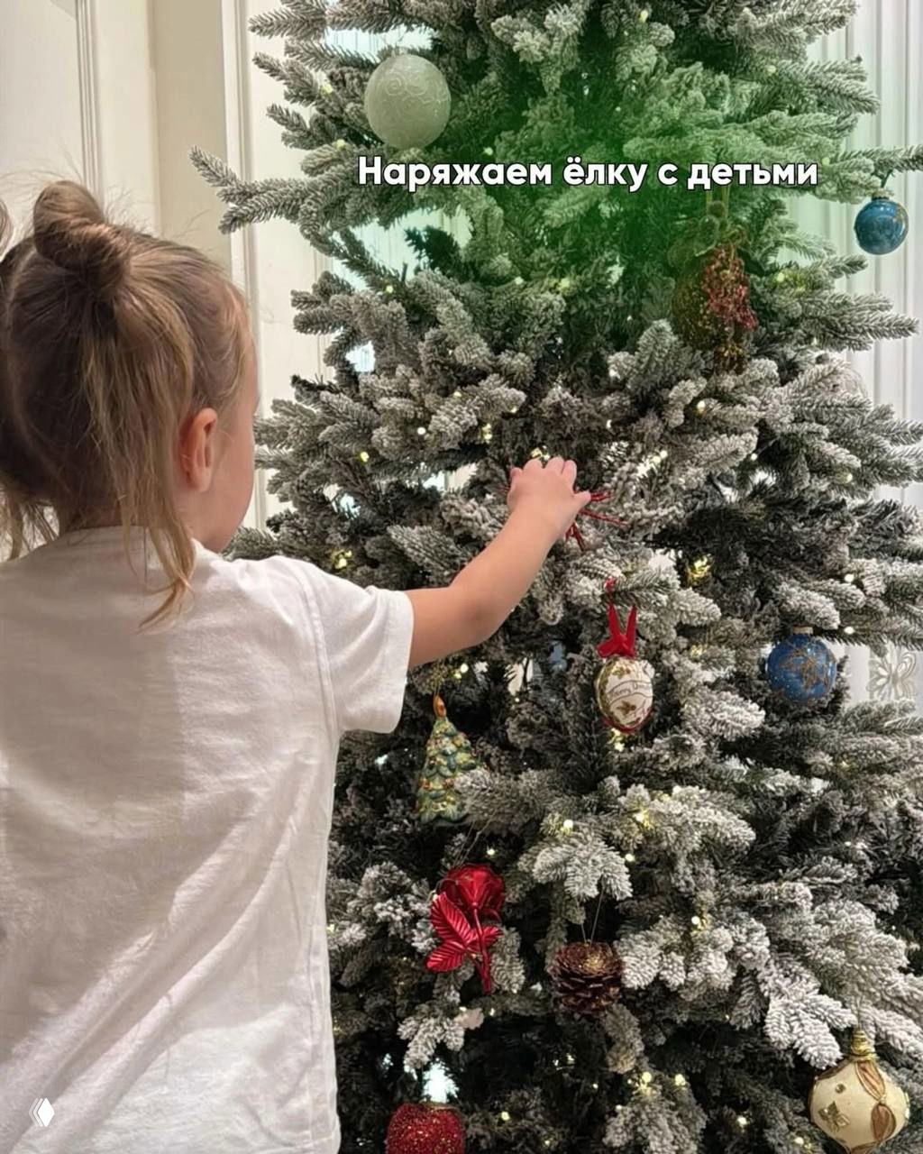 Ребёнок украшает искусственную ель с эффектом снега — кадр показывает процесс украшения ёлки вместе с детьми и семейные традиции подготовки к празднику.