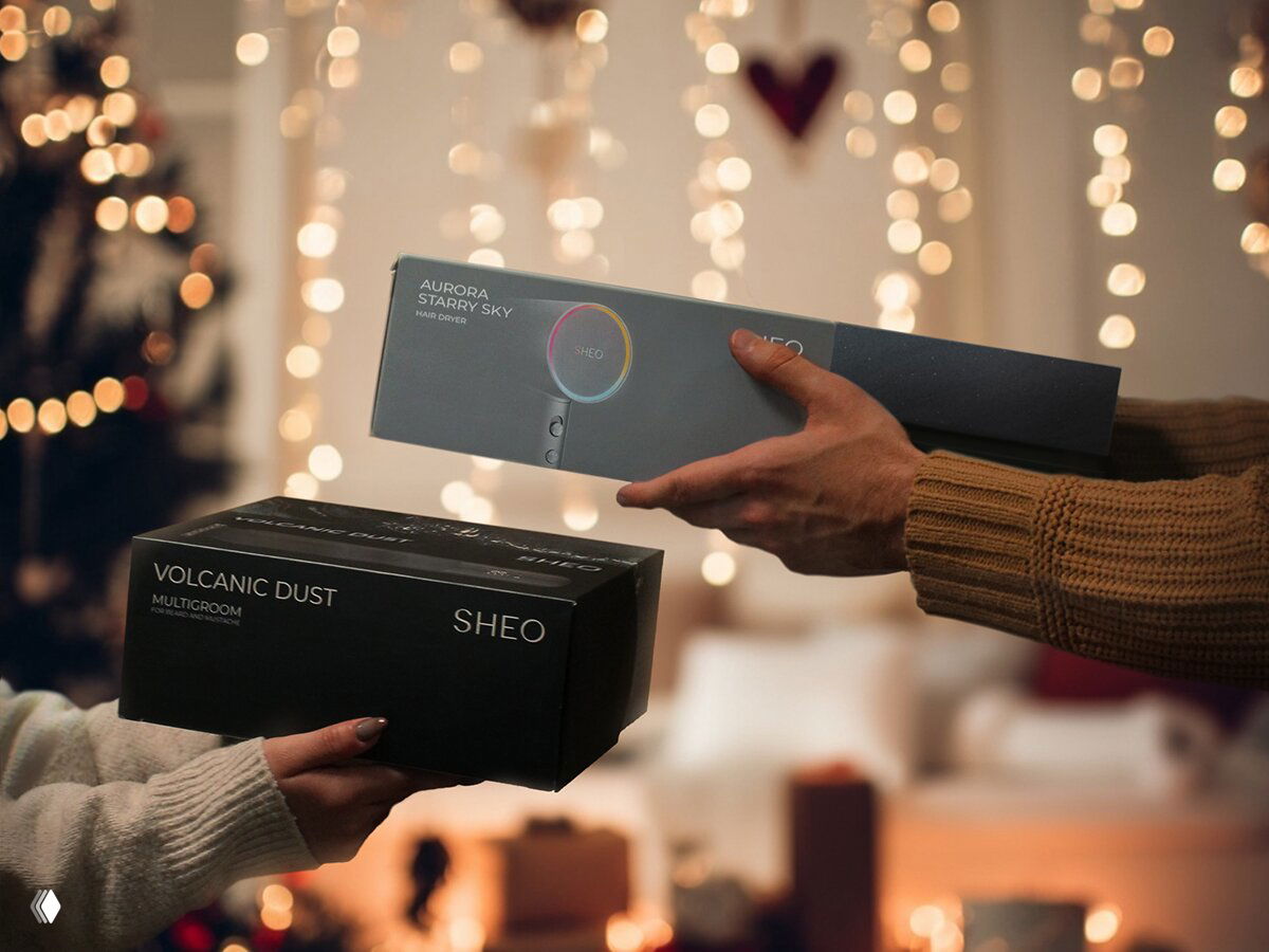 Две руки обмениваются подарочными коробками Sheo: коробка триммера Volcanic Dust и упаковка фена Aurora на размытом праздничном фоне с гирляндой.