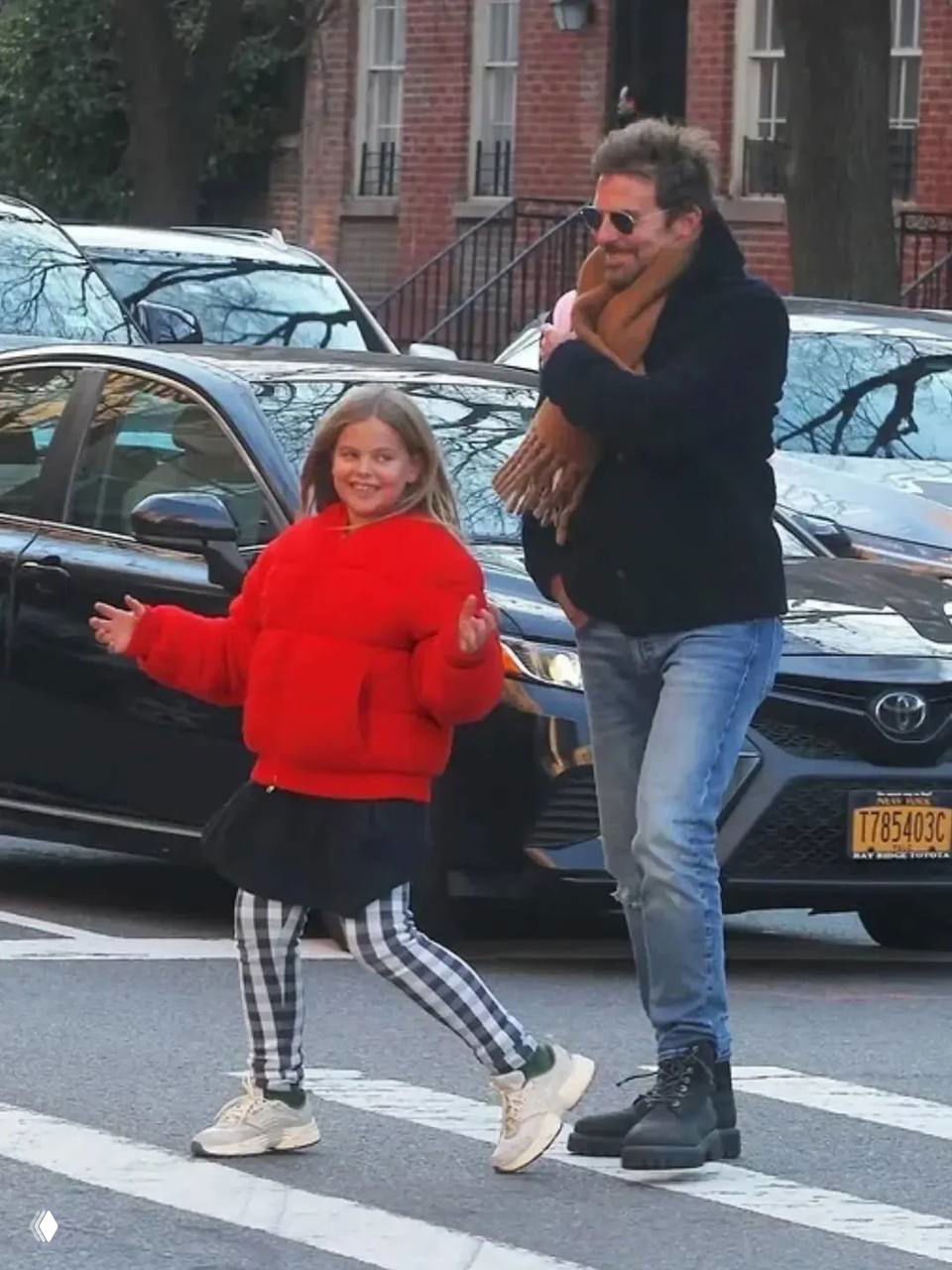 Девочка в яркой красной куртке делает шаг на переходе, рядом мужчина в джинсах и тёмной верхней одежде; снимок в уличном стиле.