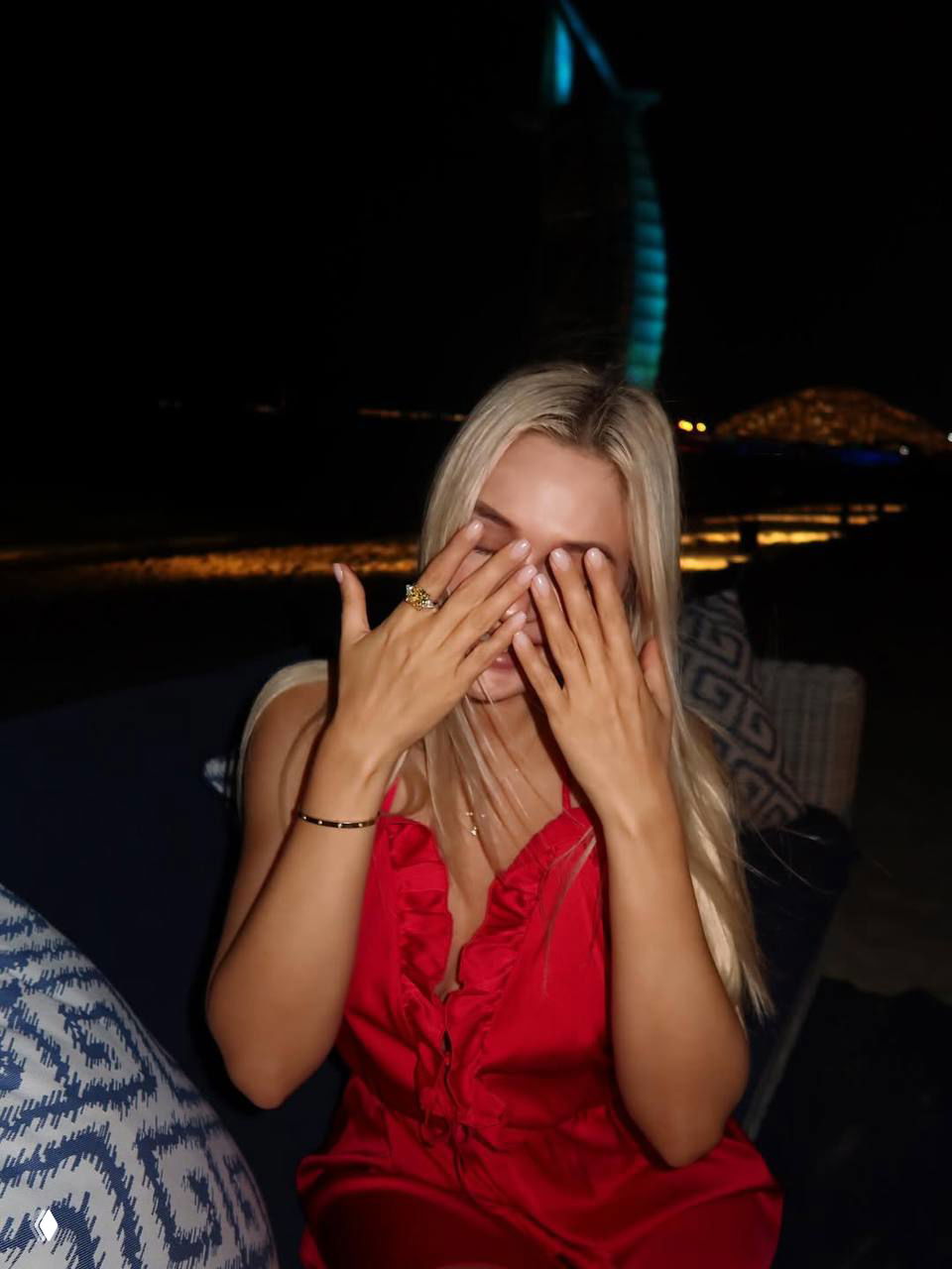 Женщина со светлыми волосами закрывает лицо руками ночью, сидя у моря; на заднем плане виден освещённый архитектурный силуэт и огни.