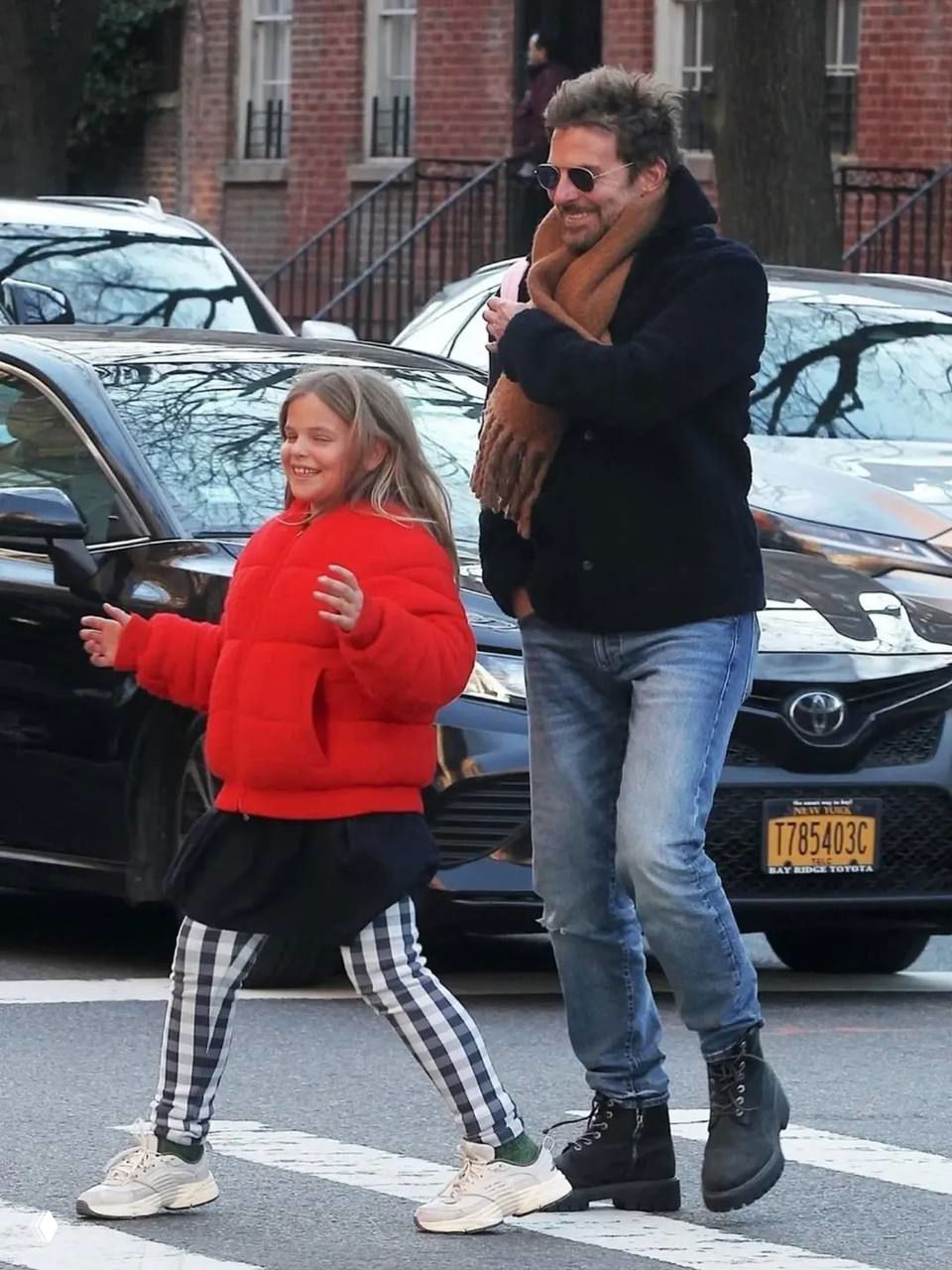 Мужчина и девочка идут по тротуару: девочка в красной пуховой куртке и в клетчатых штанах, мужчина с шарфом улыбается.