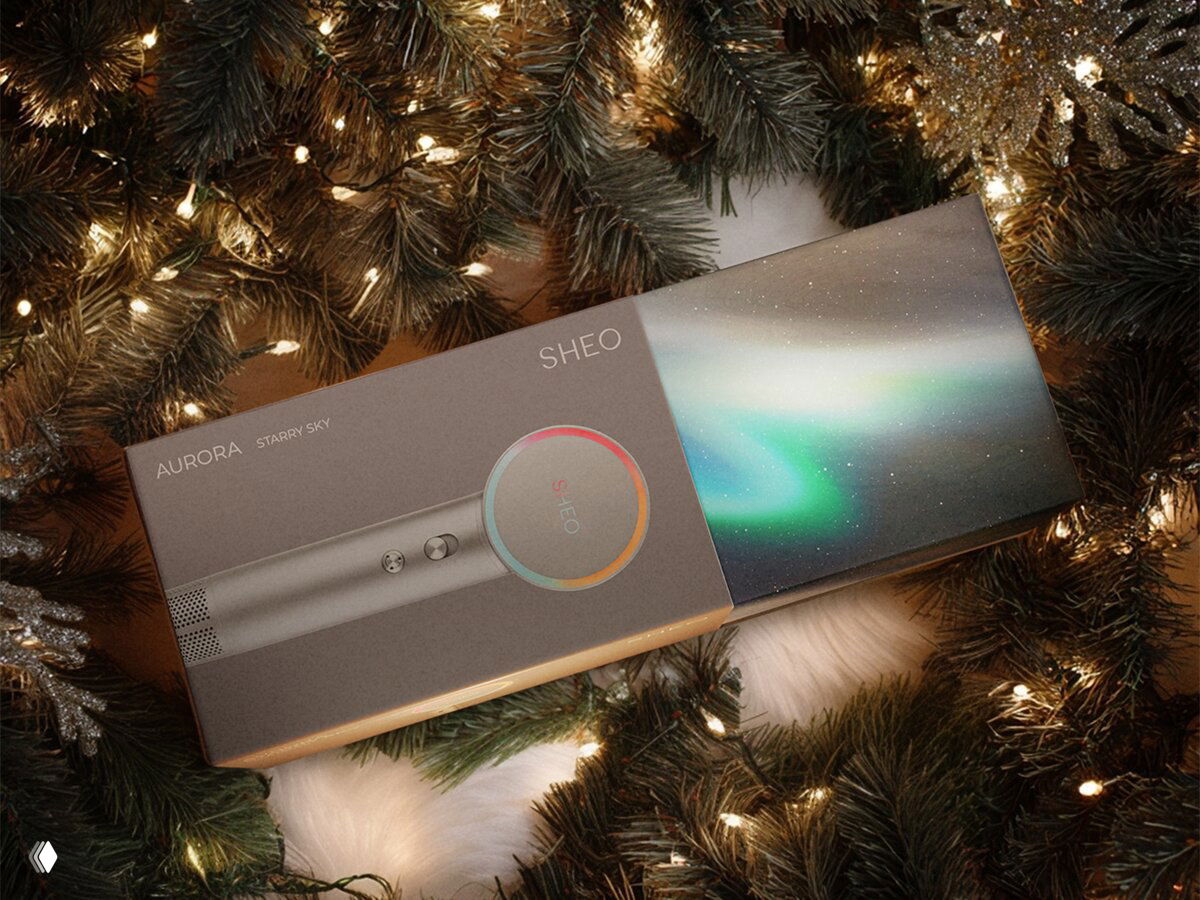 Коробка фена Sheo Aurora с изображением прибора и градиентом «северного сияния» в упаковке, лежащая среди ёлочных веток и праздничных огней.