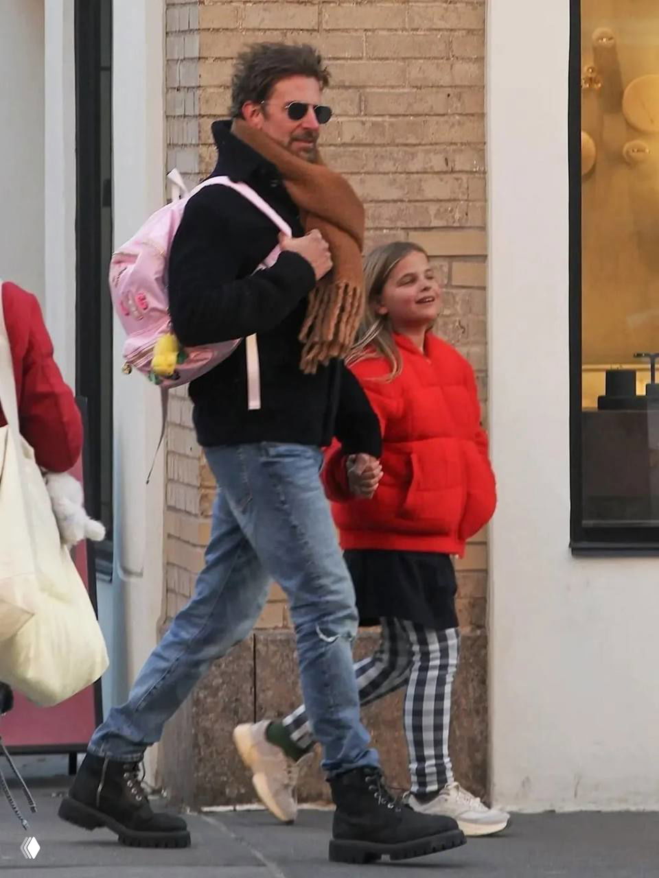 Мужчина с рюкзаком и шарфом держит за руку девочку в красной куртке; кадр снят в уличной обстановке у витрины магазина.