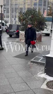 Мужчина в тёмной одежде стоит у городской ёлки на тротуаре с остатками снега; городской фон и логотип WOMAN.ru видны на фото