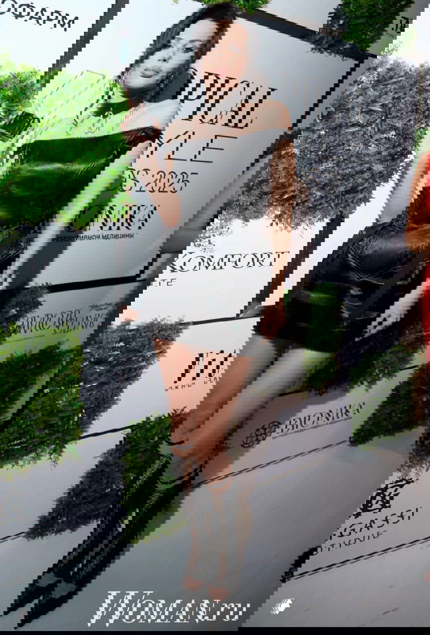 Женщина в коротком чёрном платье с чокером и высокими ботфортами держит награду на фоне бренд-волла Topical Style Awards 2025.