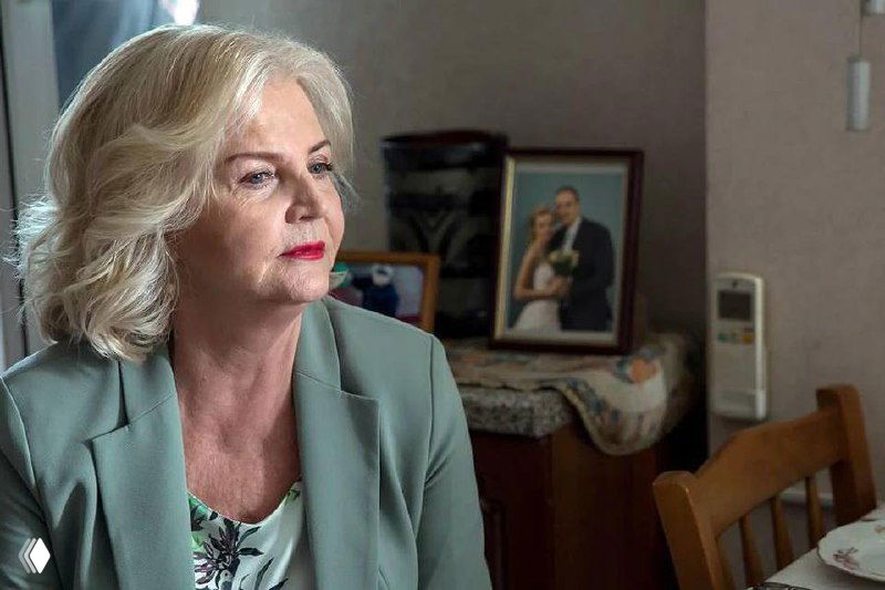 Портрет Ольги Науменко в зрелом возрасте, профиль на фоне семейных фотографий и интерьера комнаты, спокойный выразительный вид.