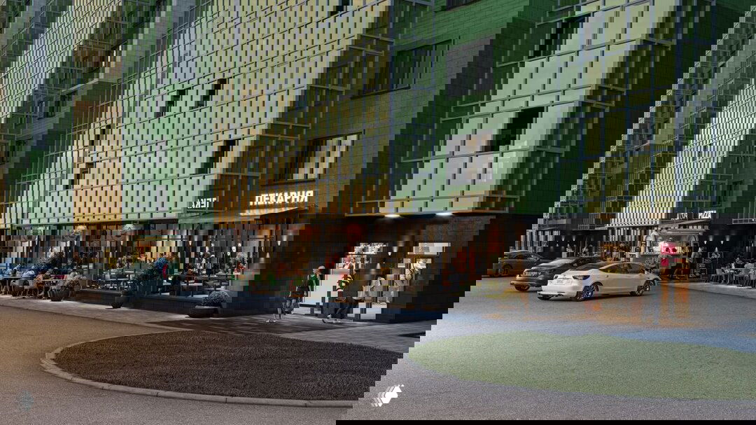 Фасад жилого комплекса «Аллея Парк»: в цоколе — кафе, пекарня и торговые помещения, парковочные места у входа, вечерняя подсветка.