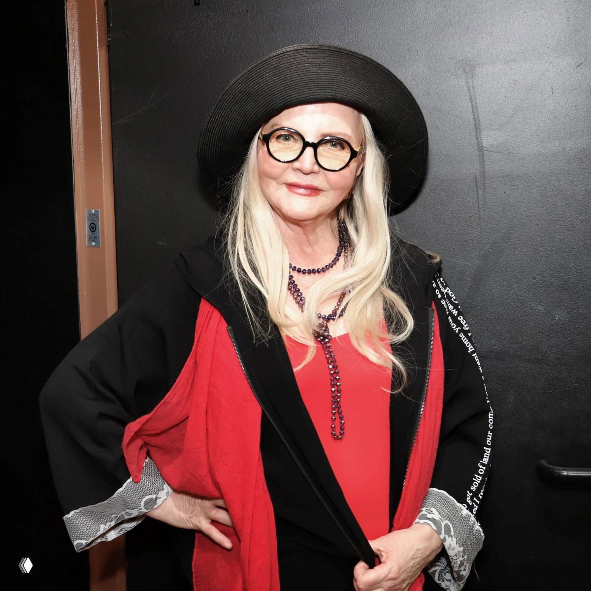 Актриса Ольга Науменко в стильном образе: светлые волосы, чёрная шляпа и очки, красно‑чёрный наряд на фоне сцены или закулисья.