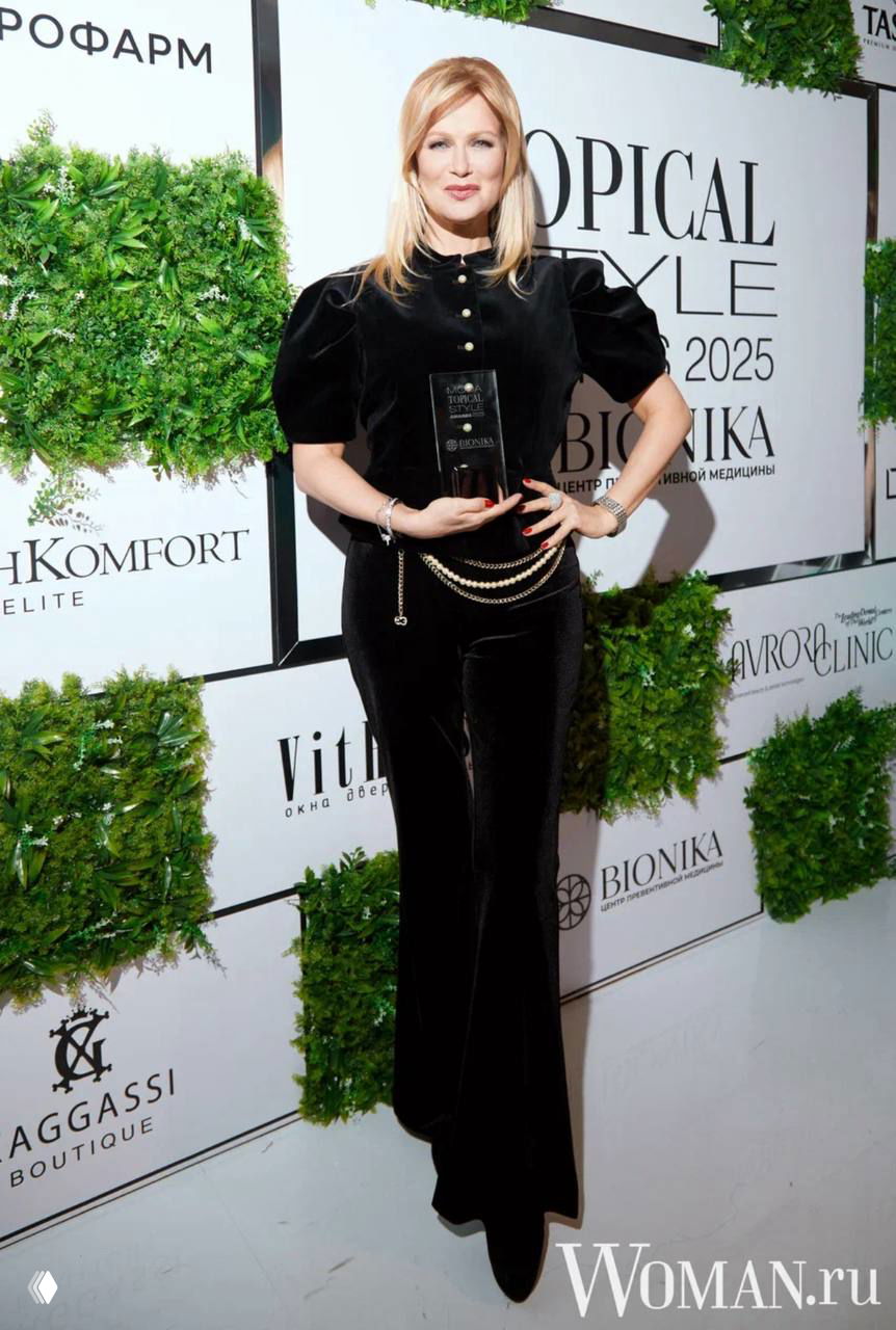 Ксения Собчак в чёрном костюме с бархатными штанами держит статуэтку премии, позируя на бренд-волле Topical Style Awards 2025.