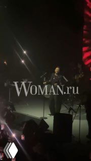 Кадр с концерта: Эмре Алтуг на сцене в другом ракурсе, подсветка зала и зрители, артист держит микрофон, логотип Woman.ru заметен
