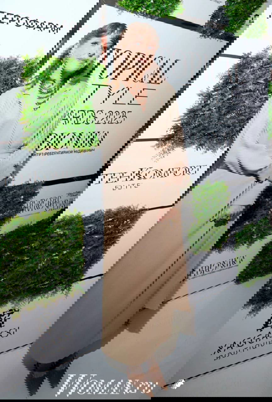 Юлия Ковальчук в светлом длинном плаще и многослойных украшениях позирует на фоне зелёных панелей и логотипов Topical Style Awards.