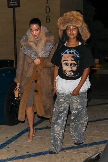 Ночной уличный снимок: подросток в тёплой шапке и свободной футболке идёт рядом со взрослой женщиной в меховом пальто, папарацци-кадр в тёмное время суток.