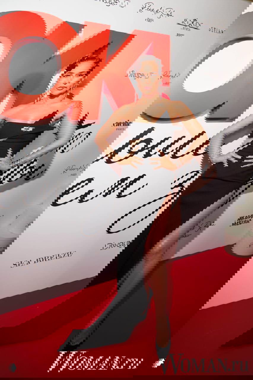 Женщина в откровенном черном вечернем платье с высоким разрезом на красной дорожке у баннера OK!, позирует фотографам, видно украшения и макияж.