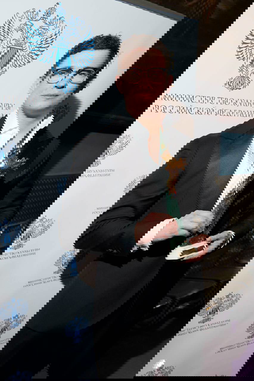 Мужчина в классическом костюме держит статуэтку премии на фоне стенда с гербом Министерства культуры, кадр с церемонии «Золотой орёл».