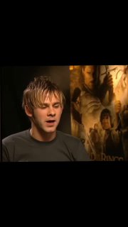 Кадр интервью: Элайджа Вуд на фоне постера «Властелина колец», средний план, нейтральная студийная обстановка и выражение лица актёра.