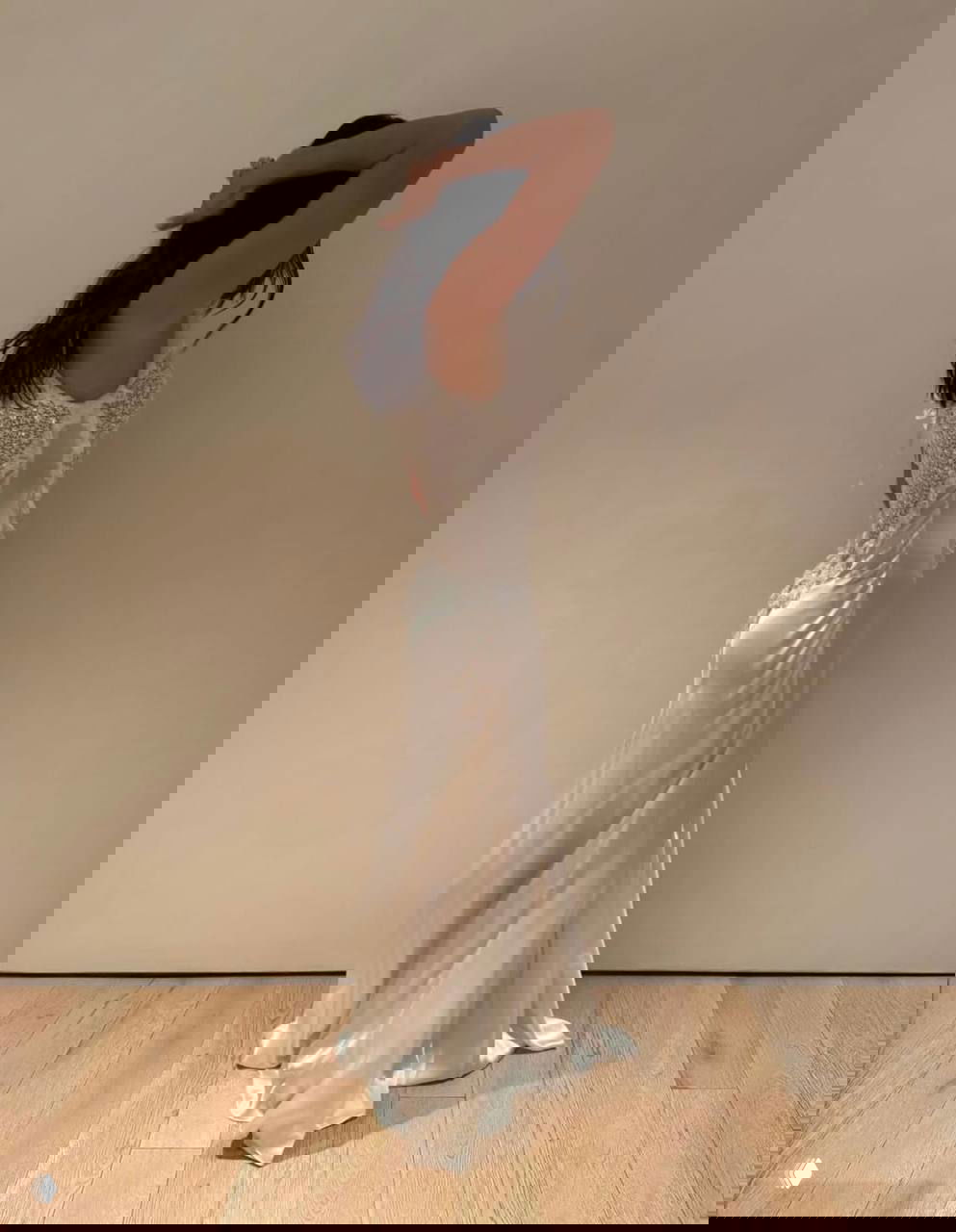 Модель в длинном белом платье снята в полный рост на фоне светлой стены; вид со стороны с акцентом на силуэт и кружевную отделку спины.