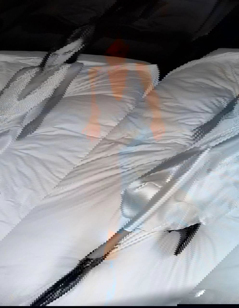 Модель в белом сатиновом платье сидит на белой кровати; крупный портретный кадр с акцентом на ткань, позу и выразительный взгляд.