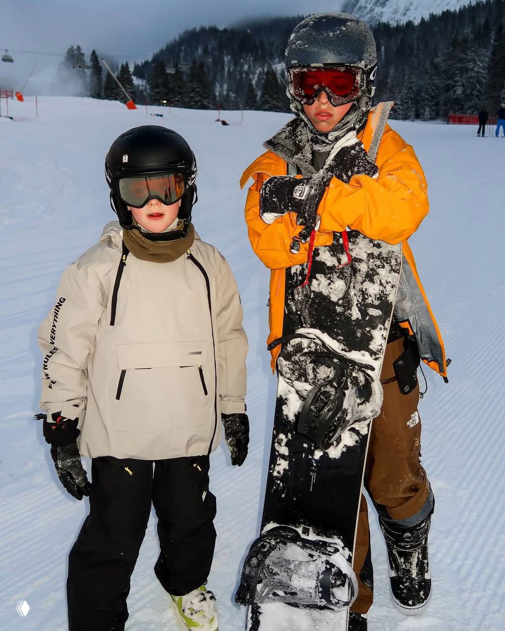 Двое детей в горнолыжной экипировке: один держит сноуборд, на заднем плане — трасса и другие отдыхающие на склоне.