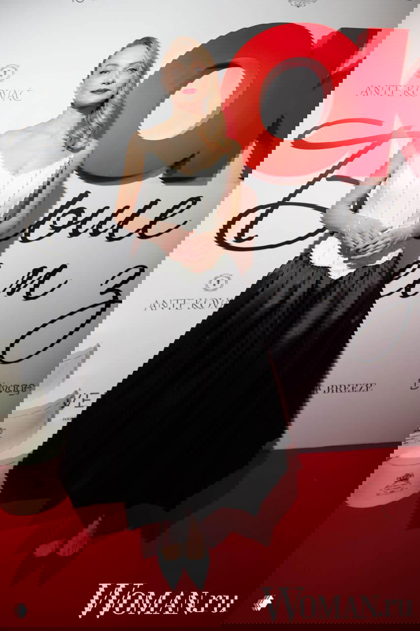 Женщина в белом корсетном верхе и пышной черной юбке стоит на красной дорожке у логотипа OK!, акцент на силуэте и классическом макияже.